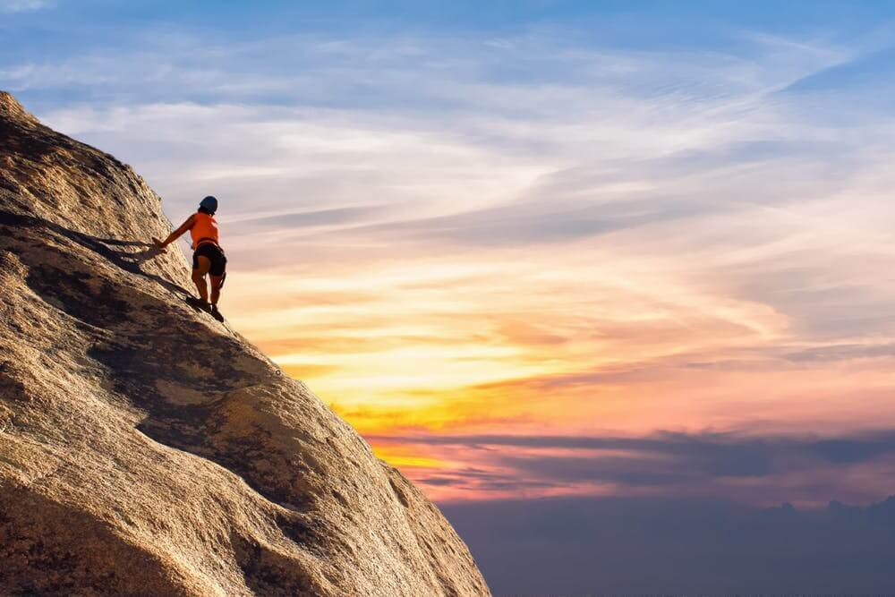 Person rock climbing at sunset with vibrant sky in the background, showcasing outdoor adventure and challenge.