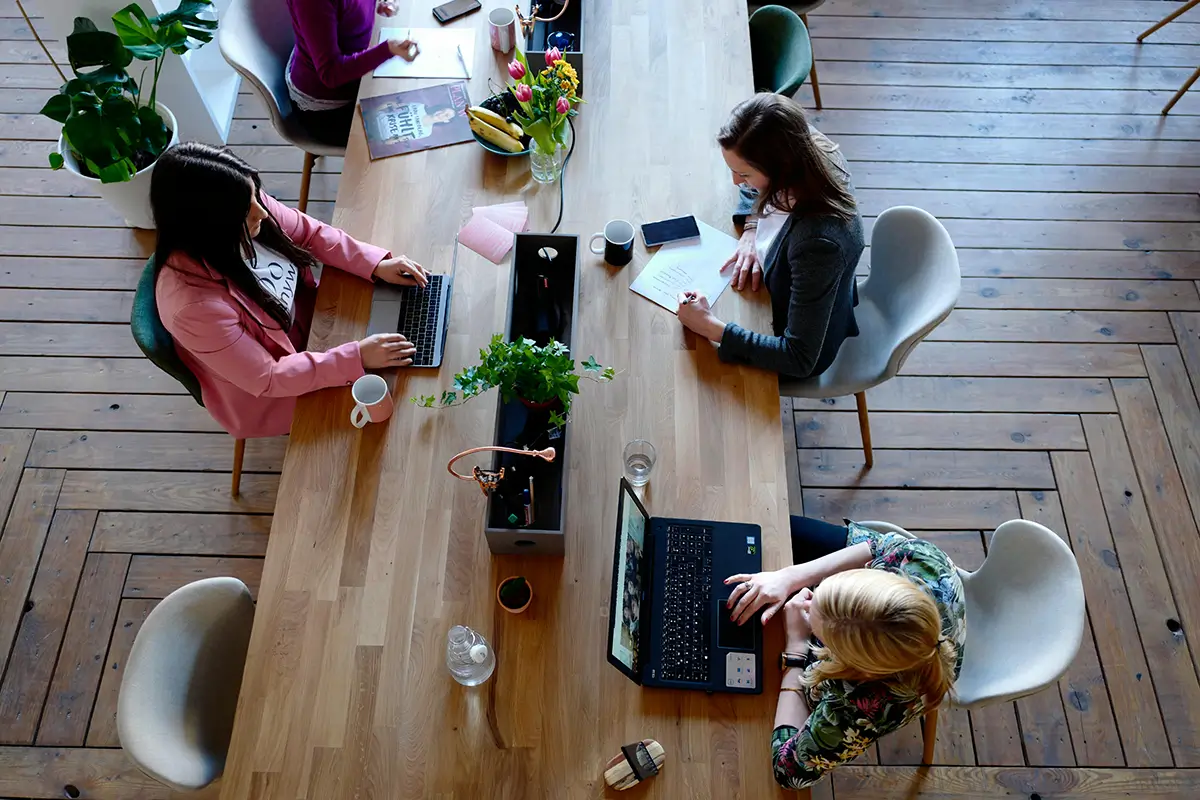 Coworkers collaborate at a wooden table with laptops, plants, and coffee in a modern office space.